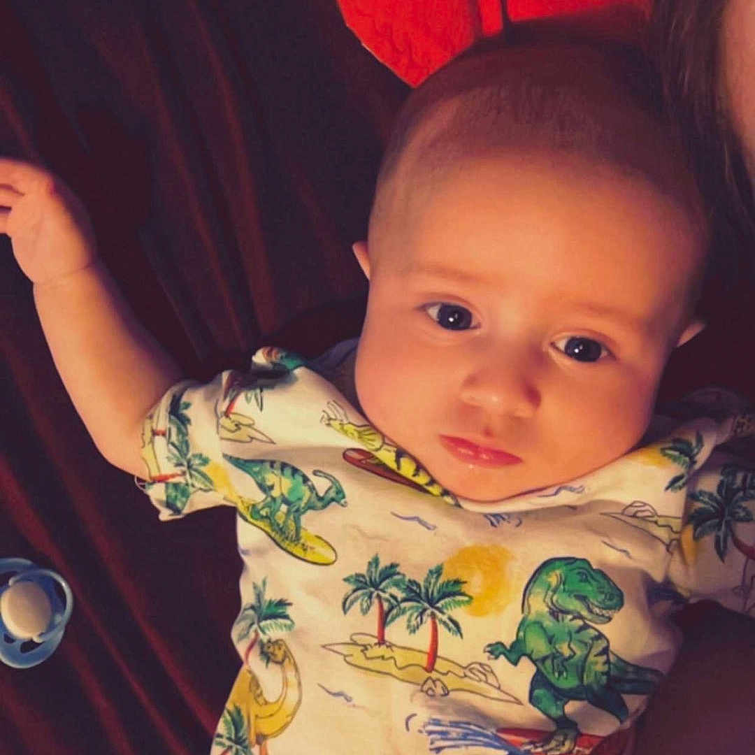 Nikko is registered to the contest to win money with this photo: baby, bed, bedding, child, clothing, cute, dinosaur, expression, face, hand, head, indoors, infant, lying_down, onesie, paciﬁer, person, portrait, sleepwear, young_child