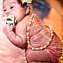 baby, infant, pacifier, flower_headband, pearl_necklace, peach_fabric, blanket, newborn, cute, sleepy, soft_lighting, portrait, closeup, skin, hand, foot, lying_down, indoors, warm_colors, adorable
