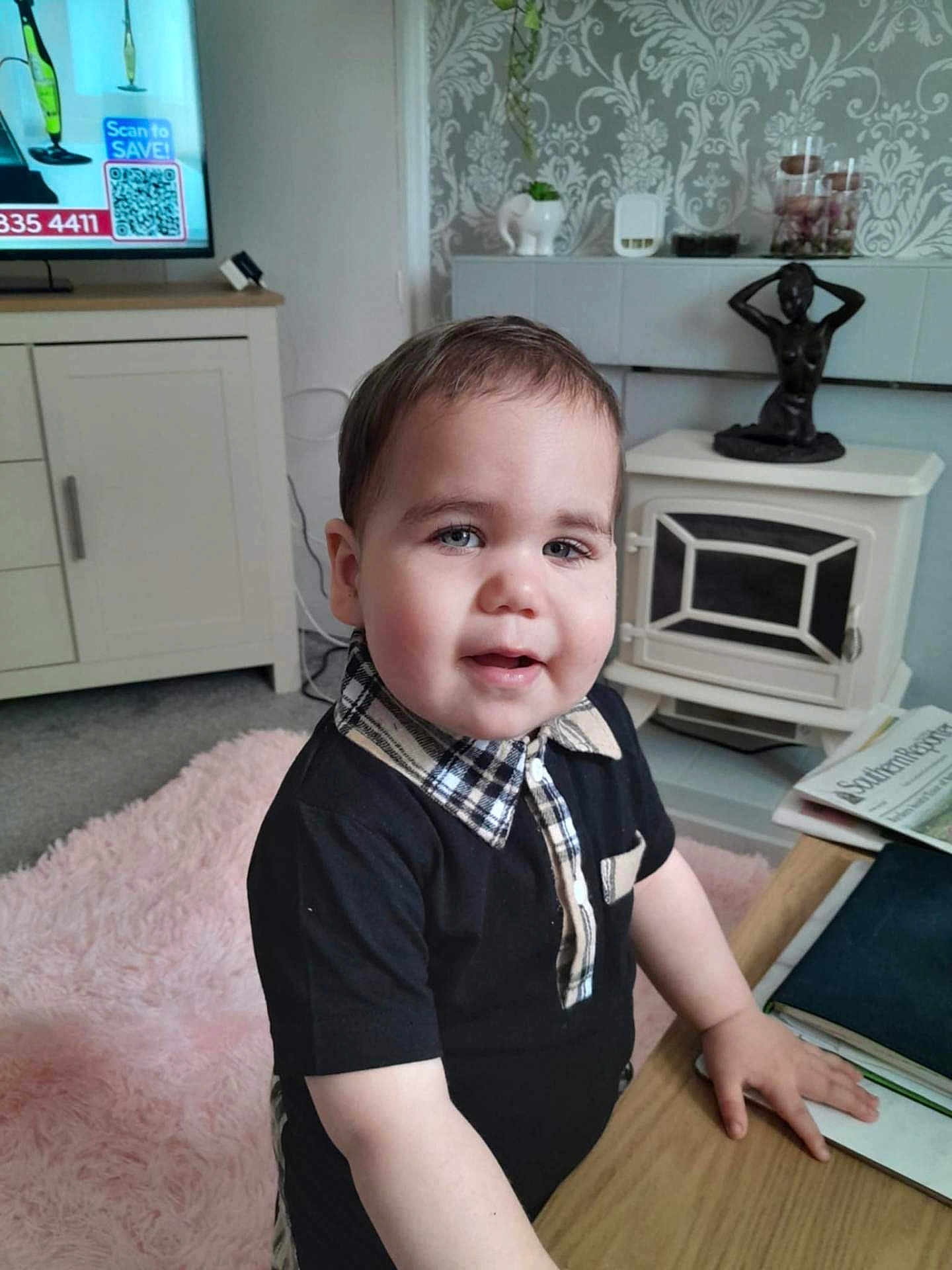 Layton is registered to the contest to win money with this photo: black_shirt, cabinet, carpet, child, cozy, curious, face, furniture, hand, home_decor, indoor, living_room, newspaper, person, plaid_collar, portrait, statue, table, toddler, young_child