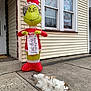 cat, fluffy_cat, inflatable, grinch, christmas, holiday_decor, sidewalk, house, window, ornaments, festive, decoration, santa_hat, naughty_list, outdoor, concrete, relaxing_cat, holiday_season, festive_lights, winter