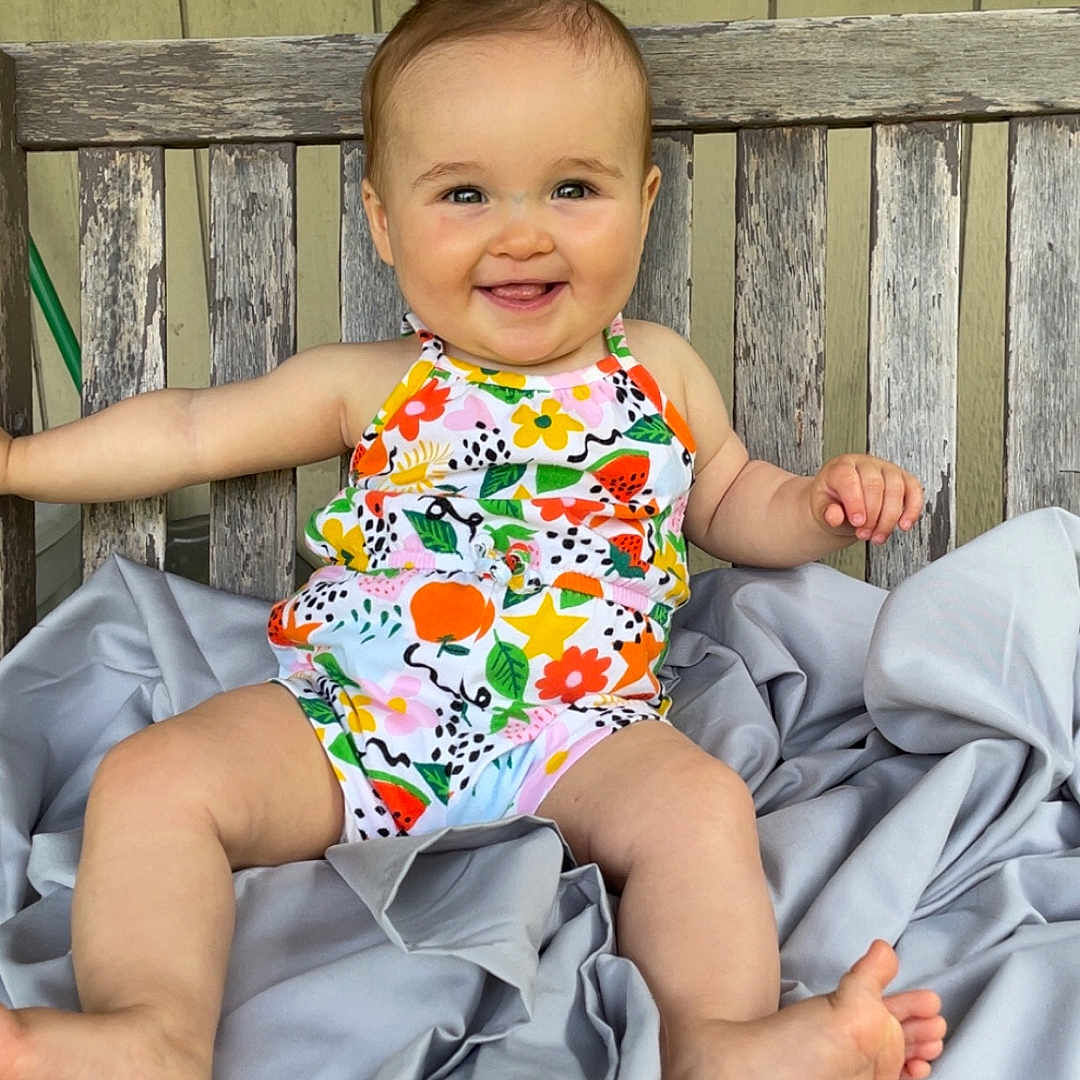 Renley is registered to the contest to win money with this photo: baby, bench, blanket, bow, child, colorful_clothing, cute, face, feet, flower_pattern, happy, infant, legs, outdoor, person, portrait, sitting, skin, smiling, wood