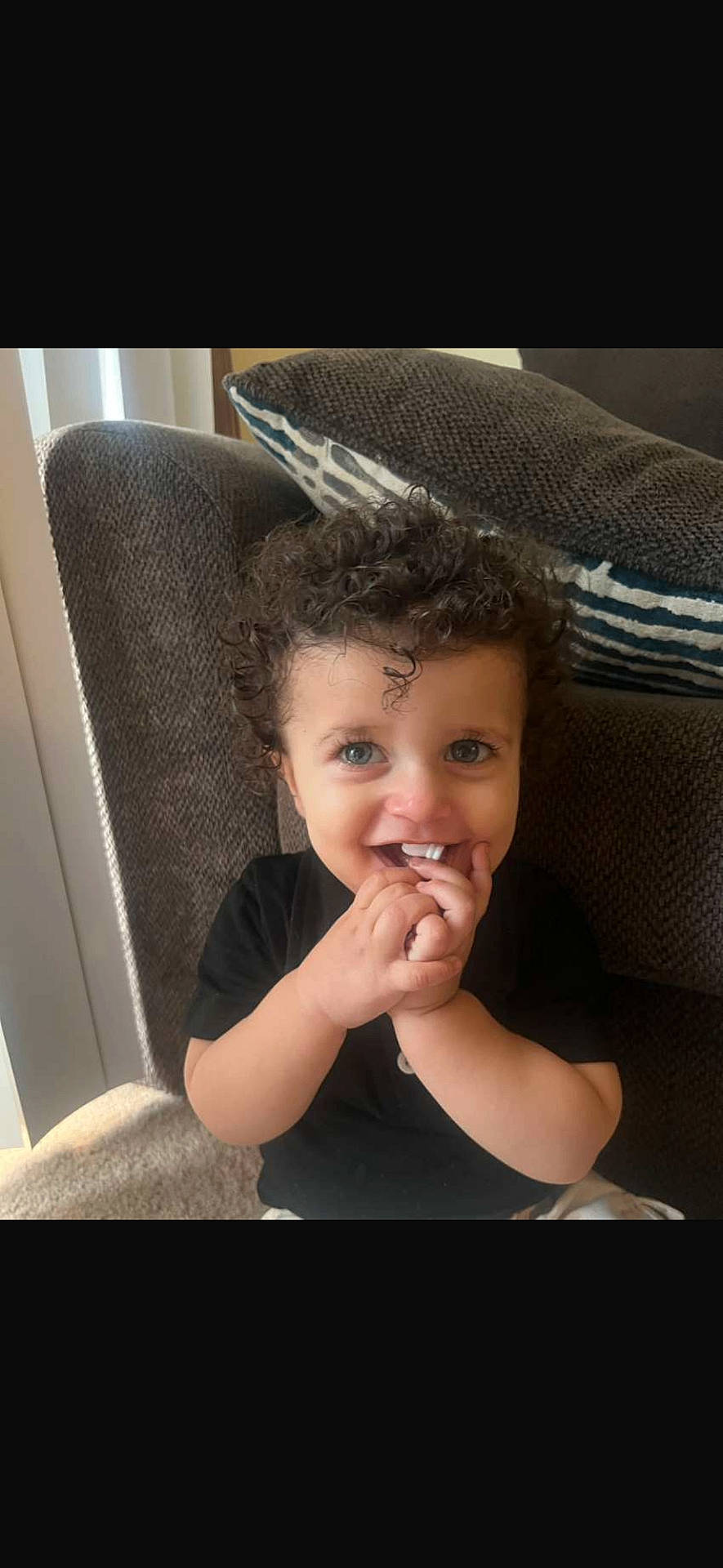 Suliman joined the competition — help win amazing prizes! black_shirt, blue_eyes, child, cozy, curly_hair, cushion, cute, fingers, happy, home, indoor, light, person, playful, portrait, smile, sofa, toddler, window_blinds