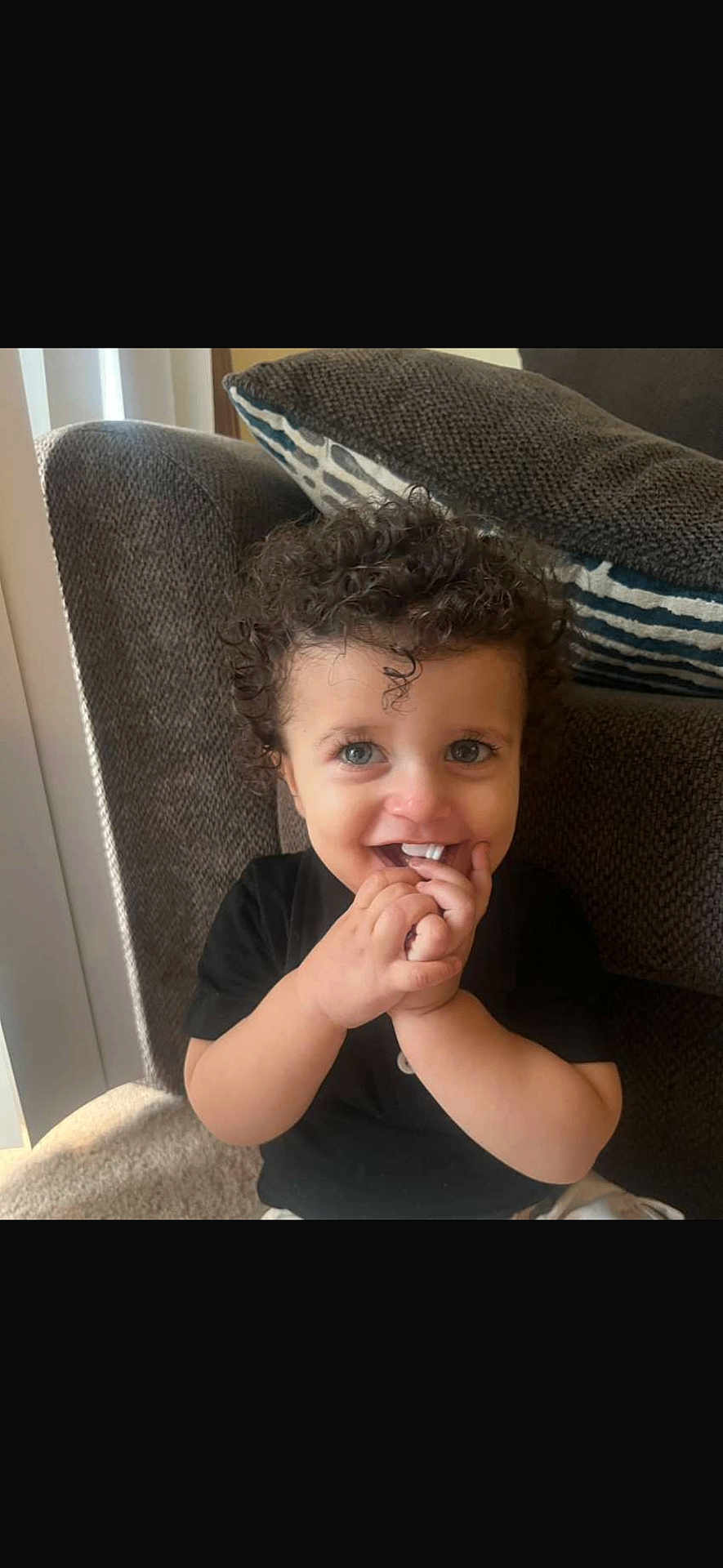 Suliman joined the competition — help win amazing prizes! toddler, child, curly_hair, smile, fingers, sofa, cushion, indoor, happy, person, black_shirt, blue_eyes, playful, cozy, home, light, window_blinds, cute, portrait