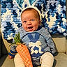 baby, child, bunny_ears, striped_sweater, carrot_toy, blue_blanket, knitted_blanket, couch, smiling, happy, indoor, footwearless, cute, headband, fabric_toy, striped_clothing, person, infant, seated, playful