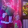 celebration, child, christmas_lights, decoration, door, face, festive, girl, green, happy, holiday, indoor, person, pink_shirt, red_clothing, santa_claus, seasonal, smile, toy, wooden_wall