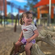 Diamond is registered to the contest to win money with this photo: child, toddler, playground, outdoor, smiling, sitting, tree_branch, sneakers, purple_shorts, light_shirt, pigtails, sunny, play_equipment, sand, happy, person, daylight, casual_clothing, park, fun