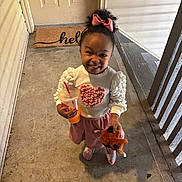 Sadie is registered to the contest to win money with this photo: child, clothing, concrete_floor, door, doormat, drink, footwear, girl, happy, heart, outdoor, person, pink_bow, porch, railing, smile, straw, stuffed_animal, sweater, toy