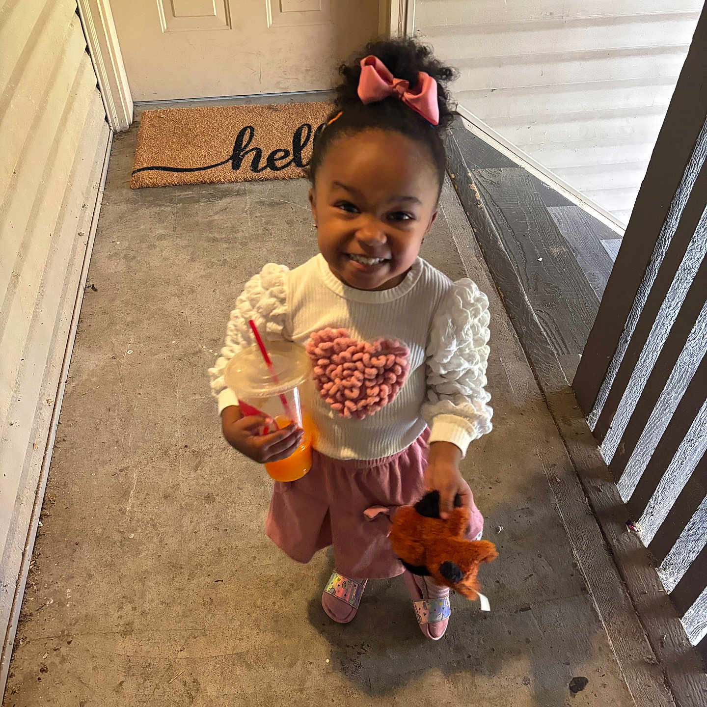 Sadie is registered to the contest to win money with this photo: child, clothing, concrete_floor, door, doormat, drink, footwear, girl, happy, heart, outdoor, person, pink_bow, porch, railing, smile, straw, stuffed_animal, sweater, toy
