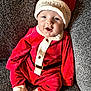 baby, child, santa_hat, christmas_outfit, red_clothing, white_trim, smiling, cute, festive, holiday, indoor, couch, fabric_texture, footwear_free, infant, happy, portrait, seated, clothing, celebration