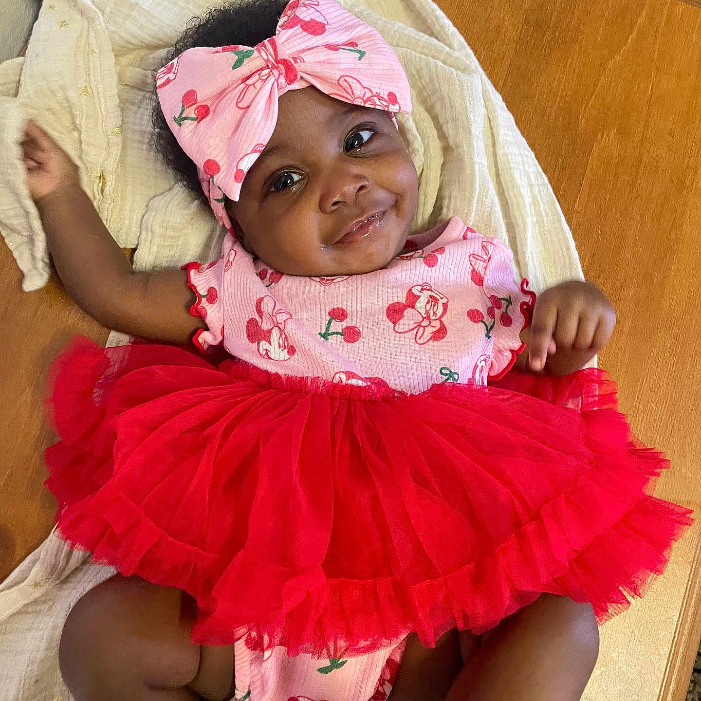 Fawlyn is registered to the contest to win money with this photo: baby, blanket, cherry, child, clothing, cute, fashion, happy, headband, indoors, infant, lying_down, person, pink, portrait, red, smile, toddler, tutu, wooden_table