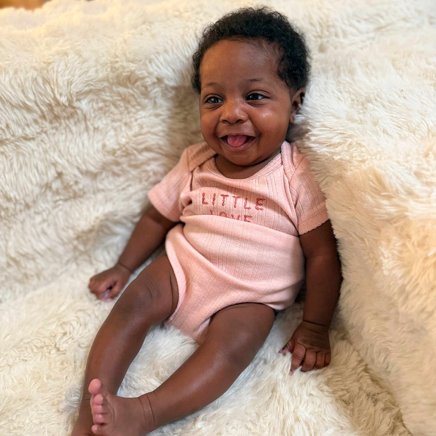 Fawlyn is registered to the contest to win money with this photo: baby, infant, smiling, pink_onesie, fluffy_blanket, happy, tongue_out, cute, child, portrait, indoor, cozy, soft_texture, sitting, young, person, skin, hair, furniture, relaxed