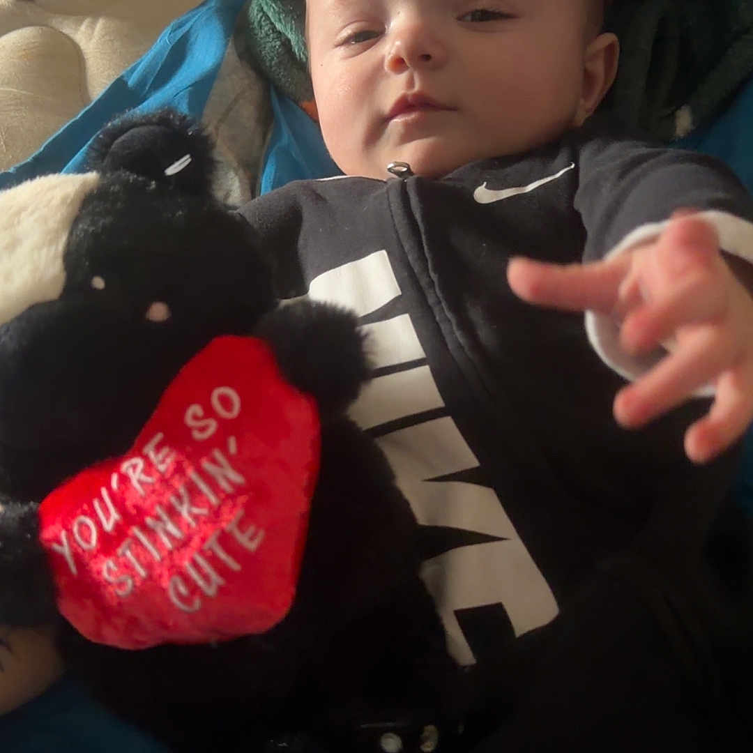 Princeston is registered to the contest to win money with this photo: baby, infant, plush_toy, skunk, heart, text_on_toy, nike_outfit, black_clothing, blanket, cozy, indoors, child, cute, soft_toy, person, smiling, lying_down, young_child, toddler, stuffed_animal
