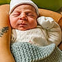 baby, newborn, sleeping, infant, white_hat, white_clothing, knitted_blanket, blue_blanket, tattoo, arm, skin, peaceful, resting, close_up, portrait, indoor, soft_light, adult_arm, baby_glove, background