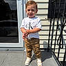 child, boy, toddler, standing, portrait, outdoor, sunlight, doorstep, house, siding, railing, stairs, window, reflection, t_shirt, pants, sneakers, fashion, smiling, daylight