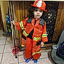 child, firefighter_costume, red_hat, indoor, curly_hair, tile_floor, toy, mop, water_dispenser, jacket, pants, shoes, reflection, wood_paneling, metal_frame, black_shirt, young_child, curious_expression, home_interior, standing
