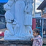 Jewlz is registered to the contest to win money with this photo: child, statue, outdoor, grass, plaid_shirt, braided_hair, stone_pedestal, fence, sky, clouds, pants, black_shoes, sculpture, person, kneeling, girl, boy, religious_statue, building, daytime