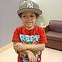 child, boy, cap, hat, red_tshirt, cartoon, gold_chain, watch, arm, couch, indoor, playful, expression, person, furniture, room, home, fashion, accessories, portrait