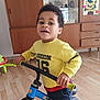child, indoor, bike, yellow_sweater, wooden_floor, cabinet, colorful_artwork, play, toy, happy, person, smiling, young_child, casual_clothing, footwear, furniture, home, curly_hair, hand, face