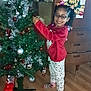 Joséphine joined the competition — help win amazing prizes! child, glasses, christmas_tree, decorations, holiday, pajamas, indoor, wooden_floor, cabinet, artwork, smile, braids, socks, festive, ornaments, tinsel, toy, flooring, person, home