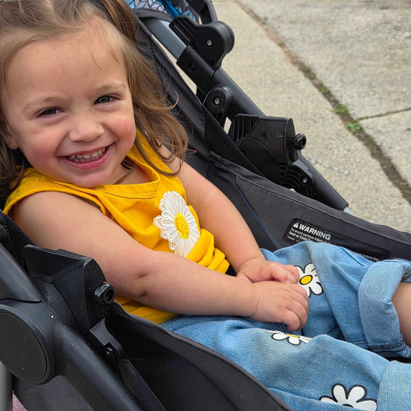 London is registered to the contest to win money with this photo: child, toddler, smiling, stroller, yellow_shirt, jeans, daisy_pattern, happy, outdoor, cute, person, casual_clothing, young_child, playful, sunny_day, seated, portrait, cheerful, baby_girl, jean_pants