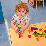 Madilynn is registered to the contest to win money with this photo: child, red_hair, curly_hair, smiling, playing, toys, building_blocks, table, classroom, books, chair, floor, educational, indoor, preschool, happy, person, colorful, learning, kid
