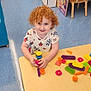 child, red_hair, curly_hair, smiling, playing, toys, building_blocks, table, classroom, books, chair, floor, educational, indoor, preschool, happy, person, colorful, learning, kid
