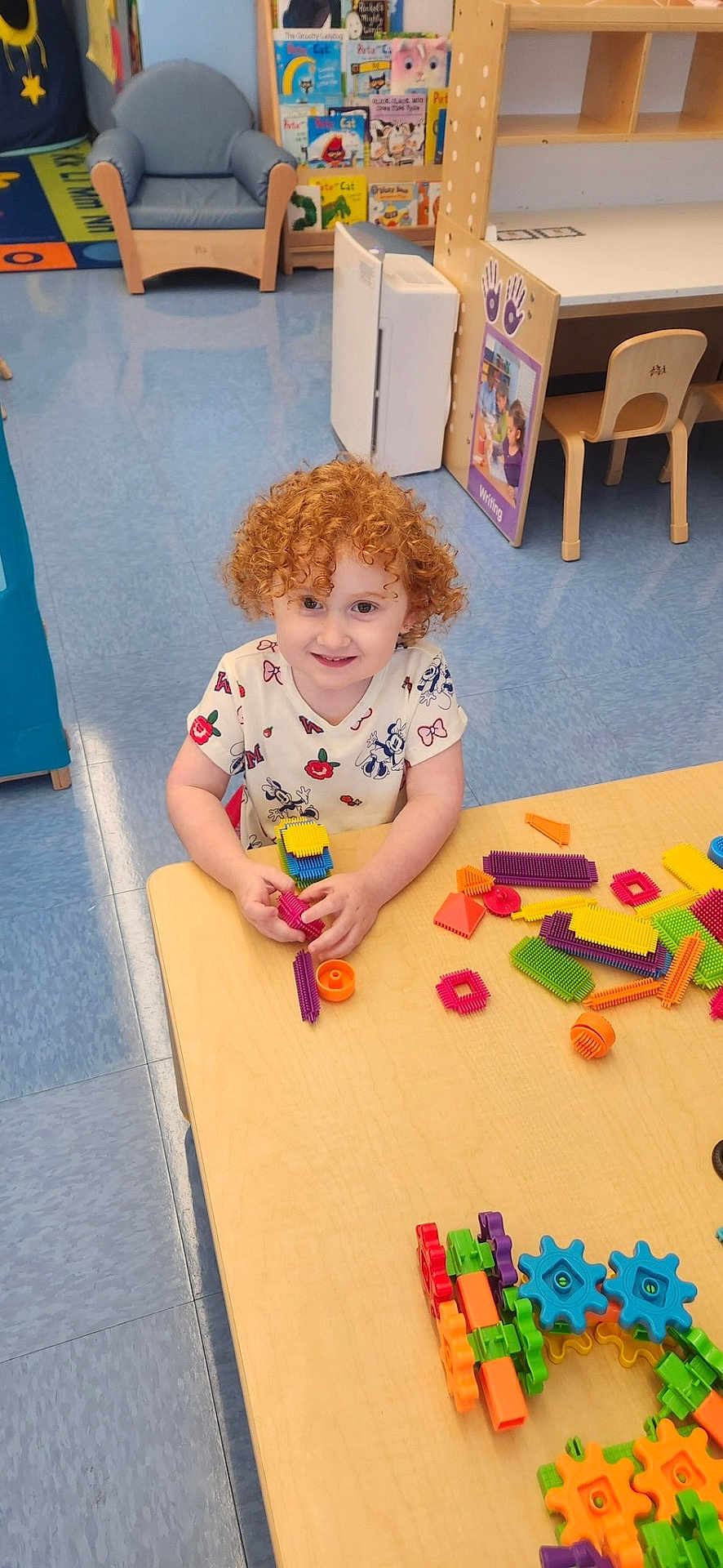 Madilynn is registered to the contest to win money with this photo: child, red_hair, curly_hair, smiling, playing, toys, building_blocks, table, classroom, books, chair, floor, educational, indoor, preschool, happy, person, colorful, learning, kid
