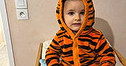 Lissandro participe au concours pour gagner de l'argent avec cette photo : bed, blanket, child, clothing, cute, eyes, hands, hoodie, indoor, onesie, orange_and_black, portrait, power_outlet, sitting, striped, tiger_onesie, toddler, towel, wall, wooden_rail