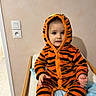 bed, blanket, child, clothing, cute, eyes, hands, hoodie, indoor, onesie, orange_and_black, portrait, power_outlet, sitting, striped, tiger_onesie, toddler, towel, wall, wooden_rail