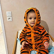 Lissandro participe au concours pour gagner de l'argent avec cette photo : bed, blanket, child, clothing, cute, eyes, hands, hoodie, indoor, onesie, orange_and_black, portrait, power_outlet, sitting, striped, tiger_onesie, toddler, towel, wall, wooden_rail