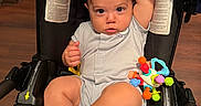 Dariel Frontela joined the competition — help win amazing prizes! baby, infant, car_seat, toy, socks, onesie, child, indoor, flooring, cute, expression, seated, colorful_toy, footwear, person, young_child, looking_up, raised_arm, black_car_seat, wooden_floor