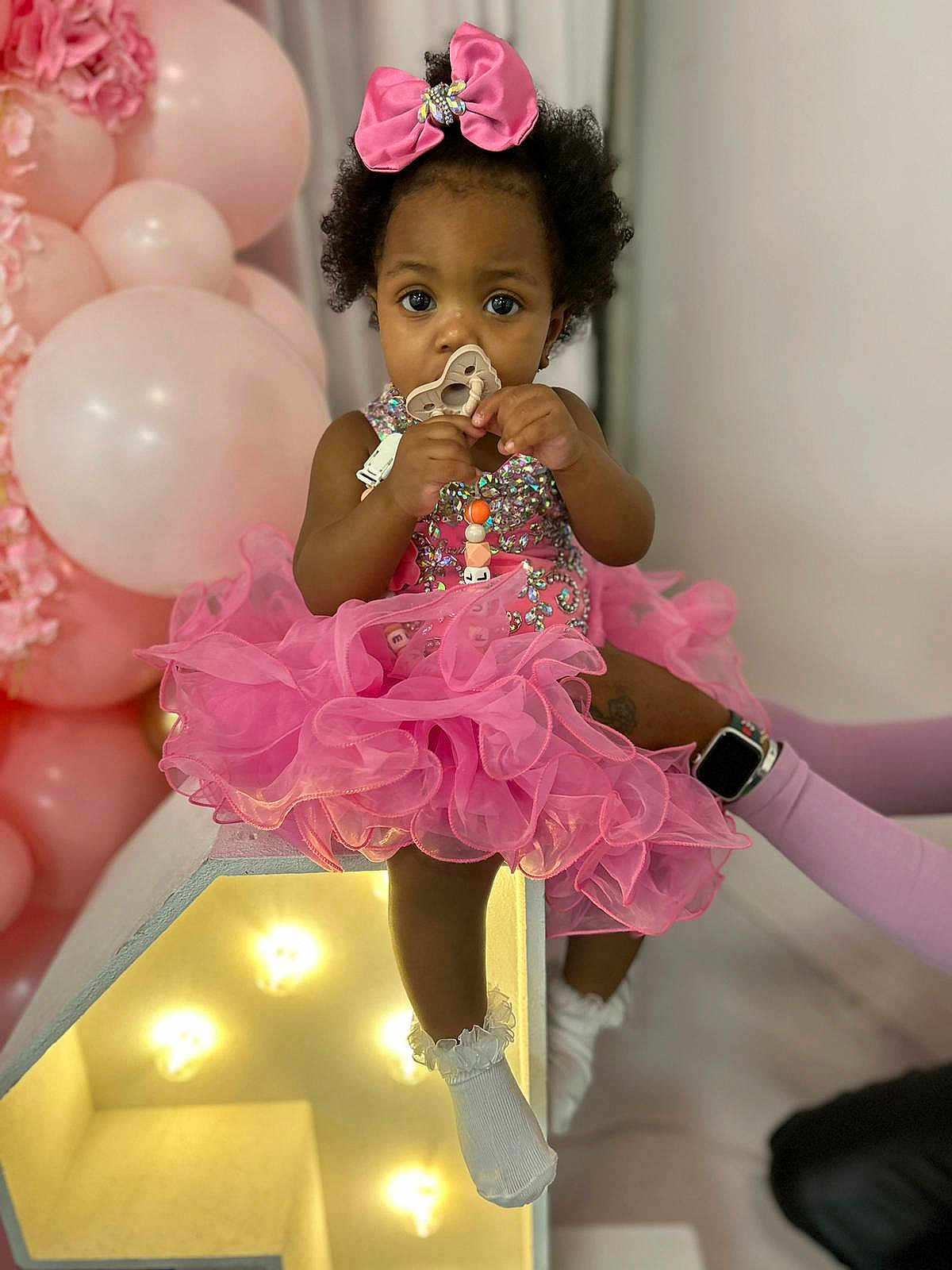 Beautiful is registered to the contest to win money with this photo: toddler, pink_dress, bow, pacifier, balloons, light_box, cute, child, celebration, party, socks, indoor, decorations, curly_hair, sparkly, festive, portrait, young_child, sitting, adorable