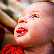 Esme is registered to the contest to win money with this photo: baby, face, tongue, red_clothing, closeup, skin, cheek, head, expression, infant, newborn, portrait, cute, child, human, person, soft_background, playful, young, smiling