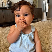 Emilia is registered to the contest to win money with this photo: baby, child, infant, romper, blue_clothing, curly_hair, sitting, indoor, carpet, rug, hand_on_face, cute, adorable, portrait, young_child, home_interior, lamp, furniture, expression, face