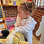 toddler, child, girl, plush_toy, rocking_horse, indoor, playroom, toy, brick_wall, white_clothing, necklace, pink_sunglasses, hairstyle, focused, furniture, carpet, colorful, window, person, cute