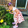 Evelyn is registered to the contest to win money with this photo: child, girl, flower, yellow_flower, purple_bow, strawberry_dress, pink_crocs, garden, green_door, brick_wall, plants, side_view, outdoor, summer, curious, nature, sunlight, concrete, casual_clothing, toddler