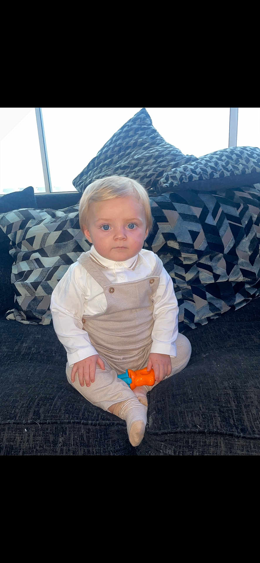 Kylo is registered to the contest to win money with this photo: toddler, child, blue_eyes, toy, couch, pillows, indoor, sitting, clothing, overalls, white_shirt, patterned, furniture, young_child, portrait, cute, serious, blond_hair, home, soft_light
