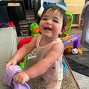 Amira joined the competition — help win amazing prizes! baby, ball, carpet, chair, child, face, furniture, happy, home, indoor, person, play_area, purple_cloth, rug, shorts, sleeveless_shirt, smiling, socks, toddler, toys