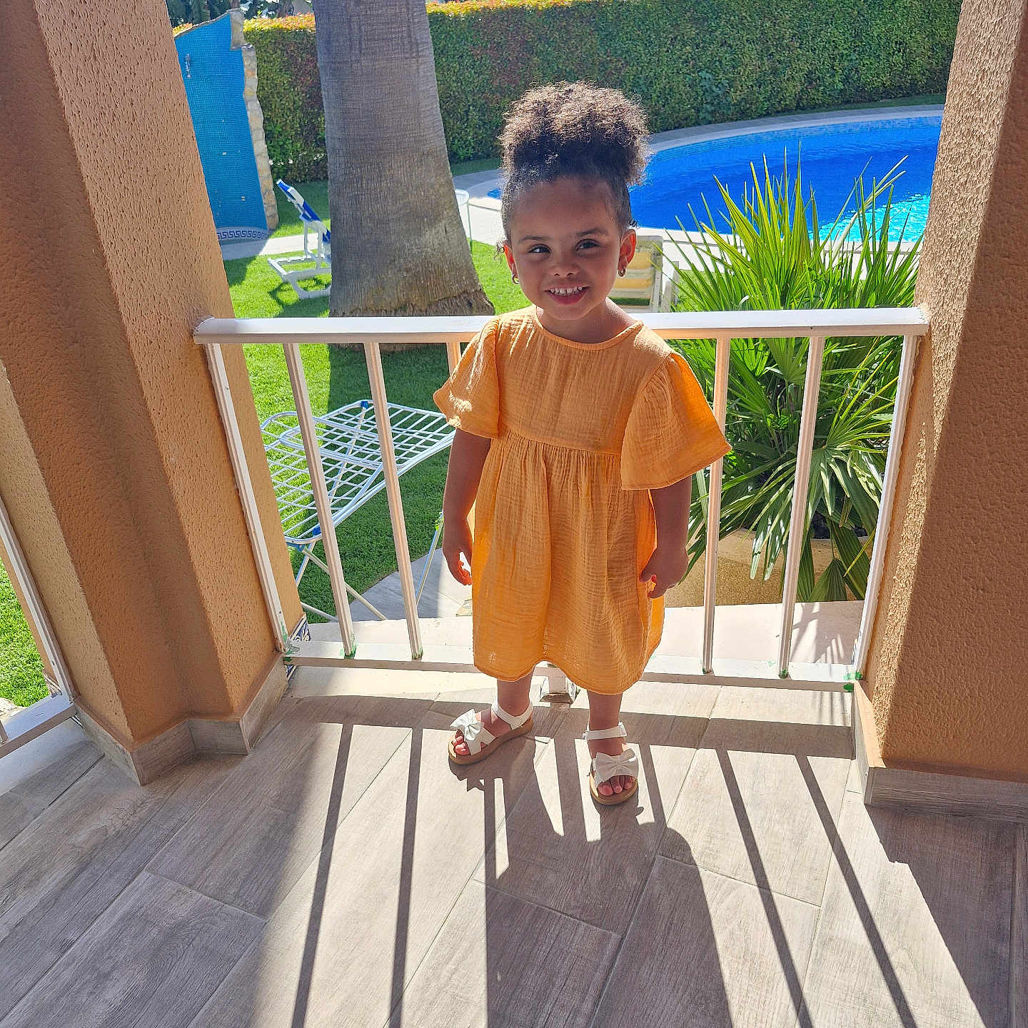 Aurora is registered to the contest to win money with this photo: balcony, child, daylight, floor, girl, greenery, happy, orange_dress, outdoor, person, plant, pool, railing, sandals, shadow, smile, summer, sunlight, tree, vacation