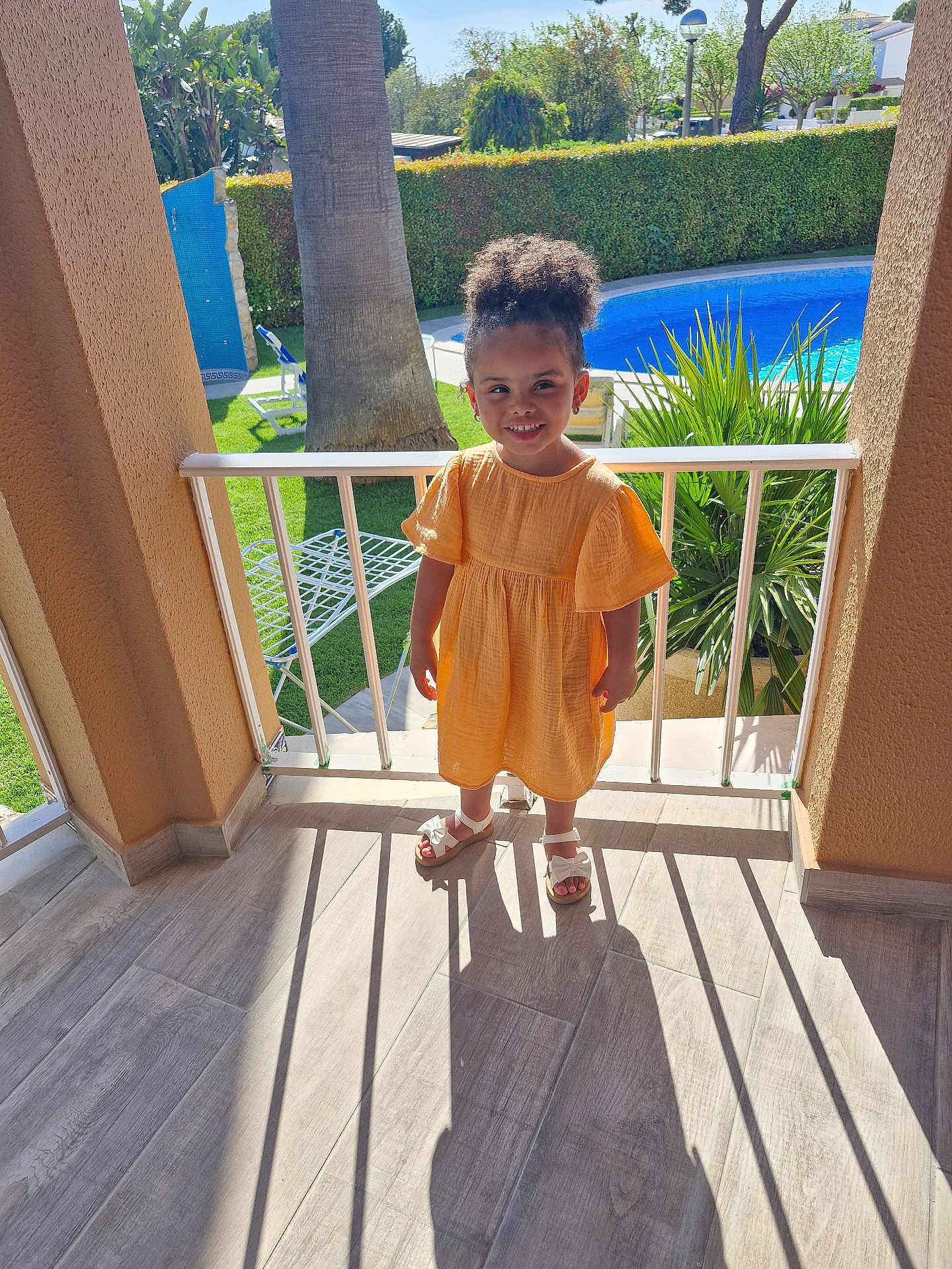 Aurora is registered to the contest to win money with this photo: child, girl, orange_dress, balcony, sunlight, shadow, pool, greenery, tree, plant, sandals, smile, outdoor, daylight, railing, floor, happy, person, summer, vacation