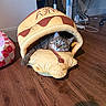 cat, tabby, pet_bed, plush, hot_dog_shape, wooden_floor, indoor, cozy, curious, paw, furniture, cables, floor, animal, cute, green_eyes, resting, home, soft, pet