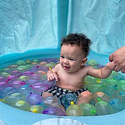 Delani joined the competition — help win amazing prizes! toddler, child, pool, water_balloons, inflatable_pool, curly_hair, smiling, holding_hand, summer, fun, water, play, outdoor, colorful, swimming, cute, baby, checkered_shorts, skin, happy