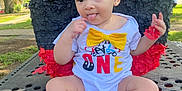 Delani joined the competition — help win amazing prizes! baby, child, outdoor, table, clothing, onesie, yellow_bow, socks, face, tongue_out, curly_hair, piñata, red, black, playful, park, daylight, earrings, cute, smiling