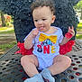 baby, child, outdoor, table, clothing, onesie, yellow_bow, socks, face, tongue_out, curly_hair, piñata, red, black, playful, park, daylight, earrings, cute, smiling