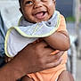 Marshall is registered to the contest to win money with this photo: baby, smiling, child, bib, orange_onesie, hands, holding, happy, portrait, infant, cute, skin, face, closeup, outdoor, blurred_background, person, toddler, joyful, expression