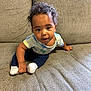 baby, child, curly_hair, couch, gray_couch, dinosaur_shirt, navy_pants, white_socks, indoors, infant, cute, portrait, sitting, looking_up, expression, hands, feet, comfortable, cozy, young_child