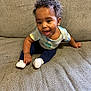 baby, child, curly_hair, dinosaur_shirt, blue_pants, white_socks, smiling, happy, indoor, couch, fabric, sitting, cute, young_child, portrait, person, home, cozy, infant, playful