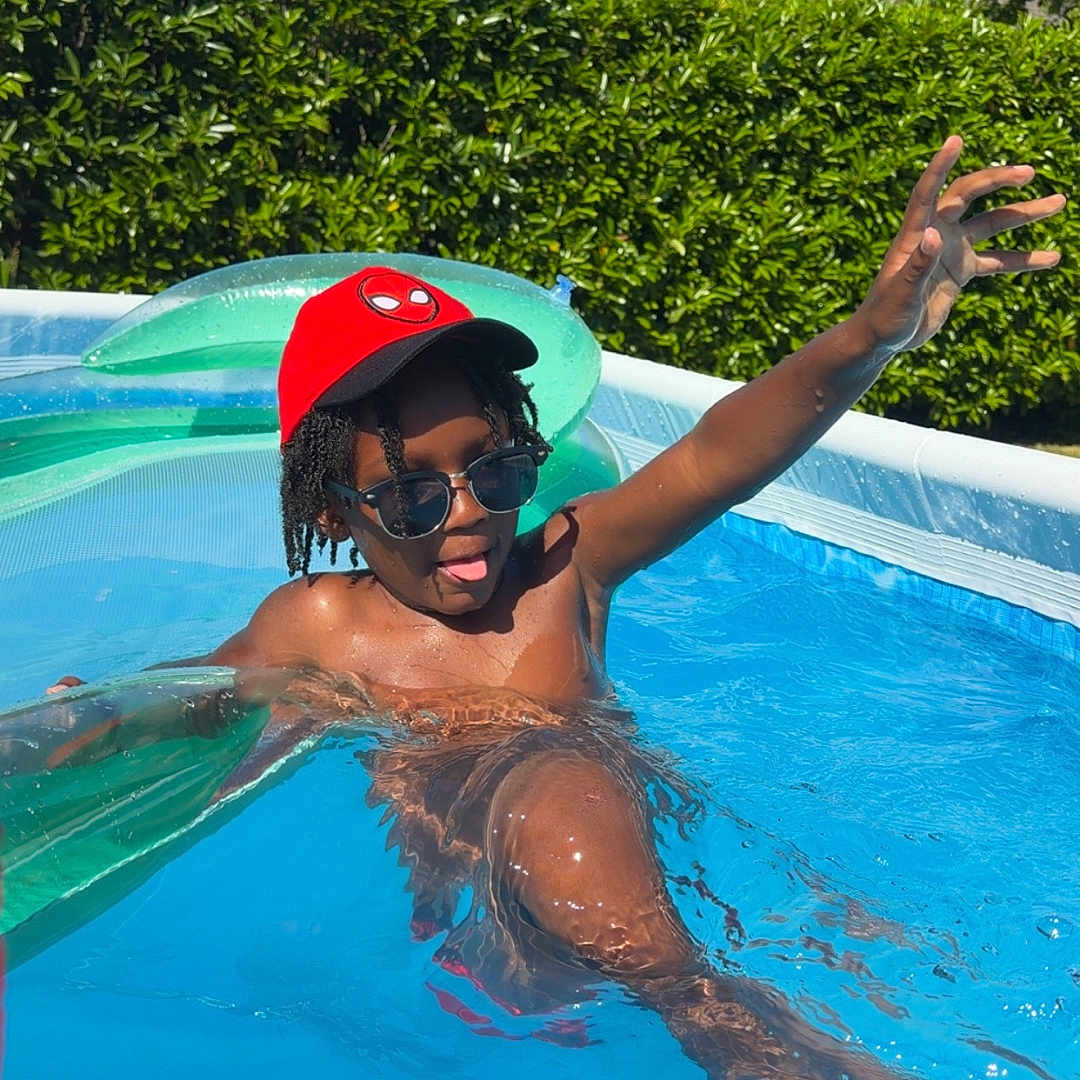 Leodrick is registered to the contest to win money with this photo: child, pool, water, sunglasses, red_cap, summer, playful, sunny, outdoor, hedge, house, inflatable, swimming, fun, daylight, person, blue_sky, relaxation, casual, child_playing