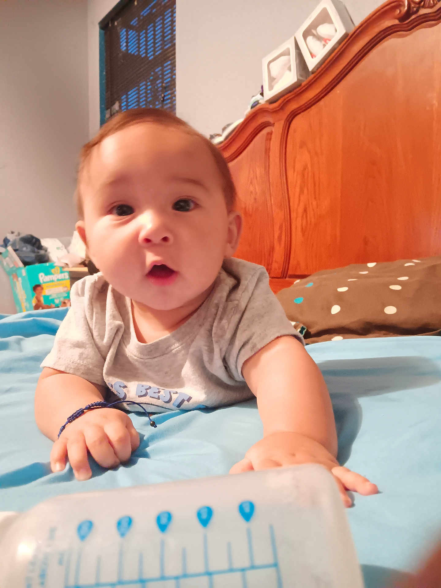 Adam is registered to the contest to win money with this photo: baby, infant, bed, bottle, wooden_headboard, pillow, blanket, room, diapers, window, curtains, clothing, hand, face, person, indoor, blue, brown, expression, child