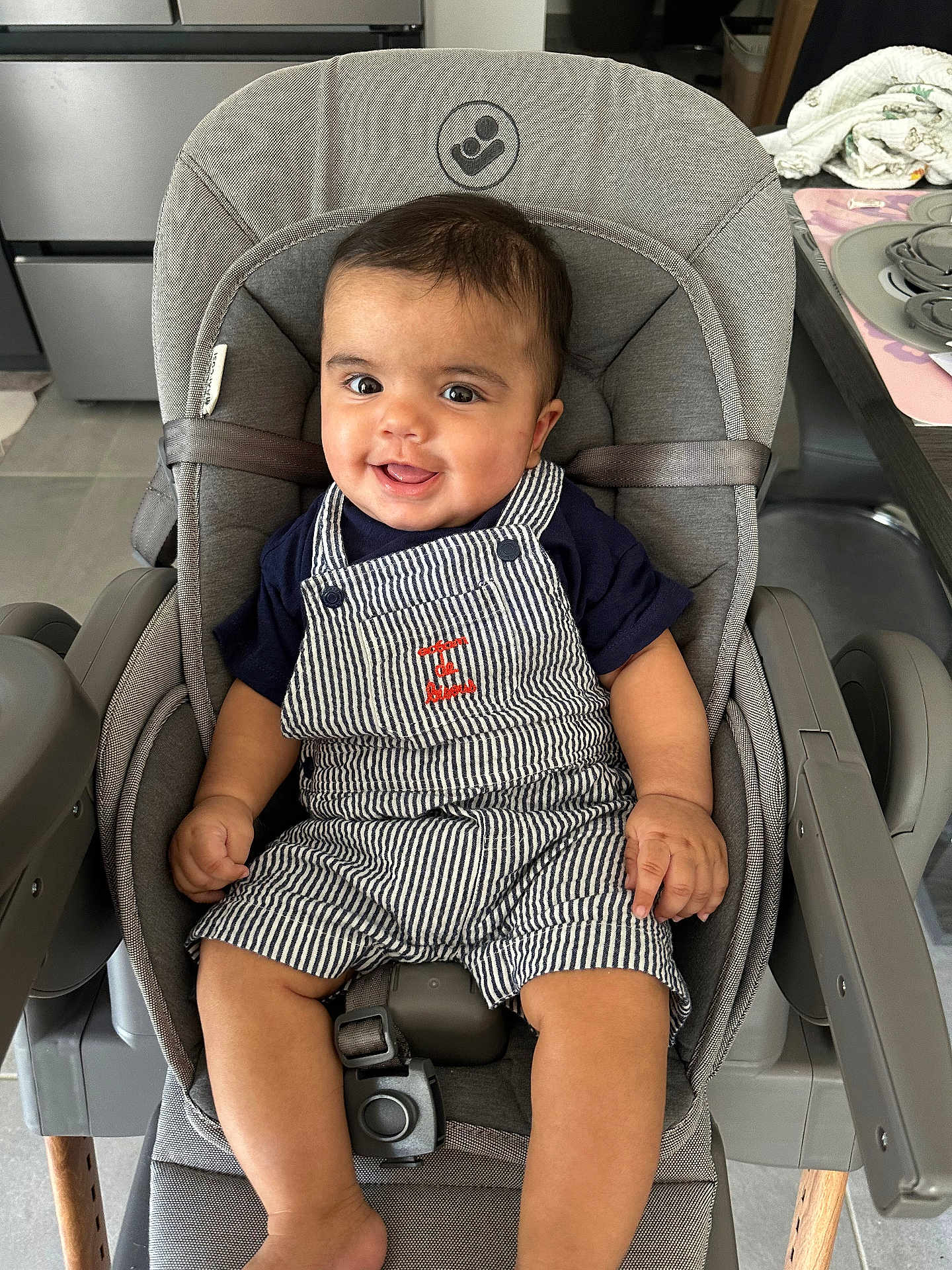 Maël a rejoint le concours — aidez-le/la à gagner de superbes lots ! baby, child, smiling, high_chair, overalls, striped_clothing, navy_shirt, indoor, kitchen, table, cloth, seat_belt, happy, face, person, infant, wooden_legs, modern, cushion, furniture